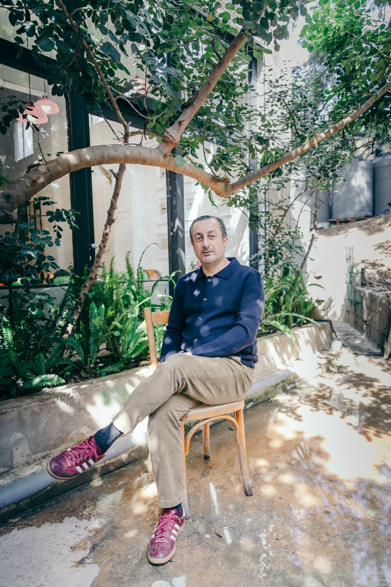 Kamal Mouzawak sits on a wooden chair in a lush garden courtyard with plants and vines overhead