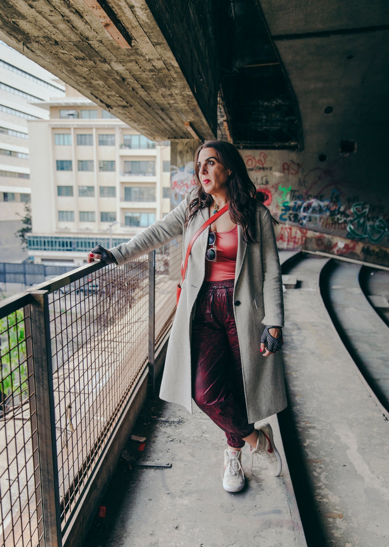 Rana Salam stands on a concrete walkway under a bridge, wearing a long gray coat and looking upward