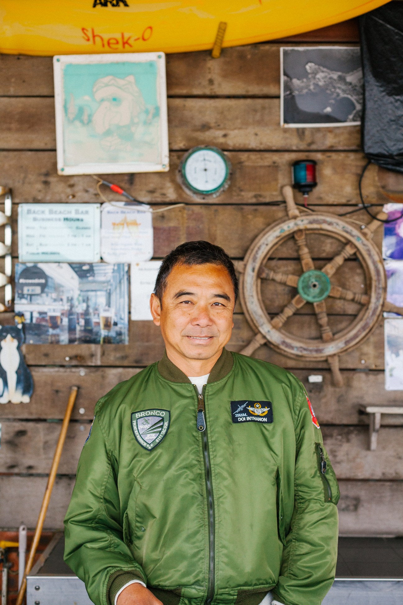 Ben Lau, owner of Ben's Back Beach Bar in Shek O, Hong Kong, wearing a green jacket in front of a wooden wall decorated with nautical items.