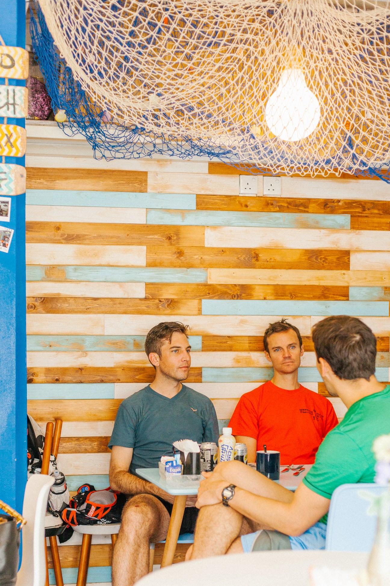 Three men having drinks at a casual beachside café with colorful wooden wall and fishing nets.