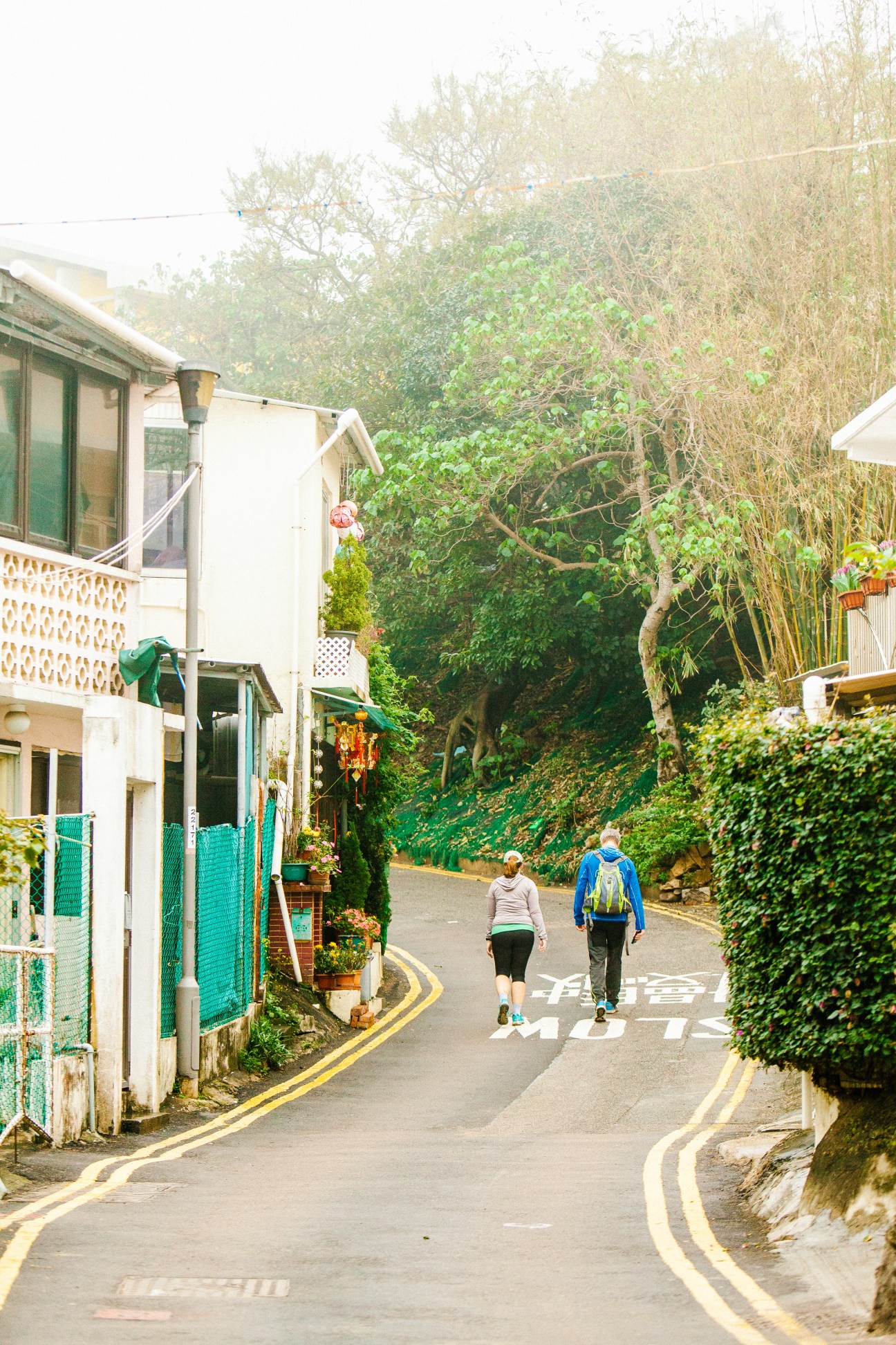 Two people jogging up a narrow residential street lined with white houses and lush green vegetation in Shek O, Hong Kong.