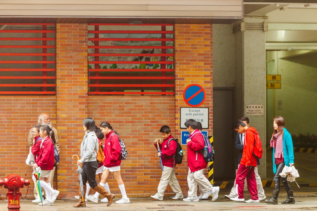 Students in red and pink school uniforms walking past a brick building with red horizontal bars in Hong Kong's Tai Hang neighborhood.