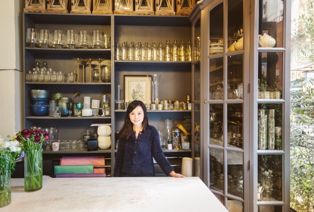 Joyce Li, director of JF Floral Couture, stands in front of shelving displaying glassware and home décor items in her shop.