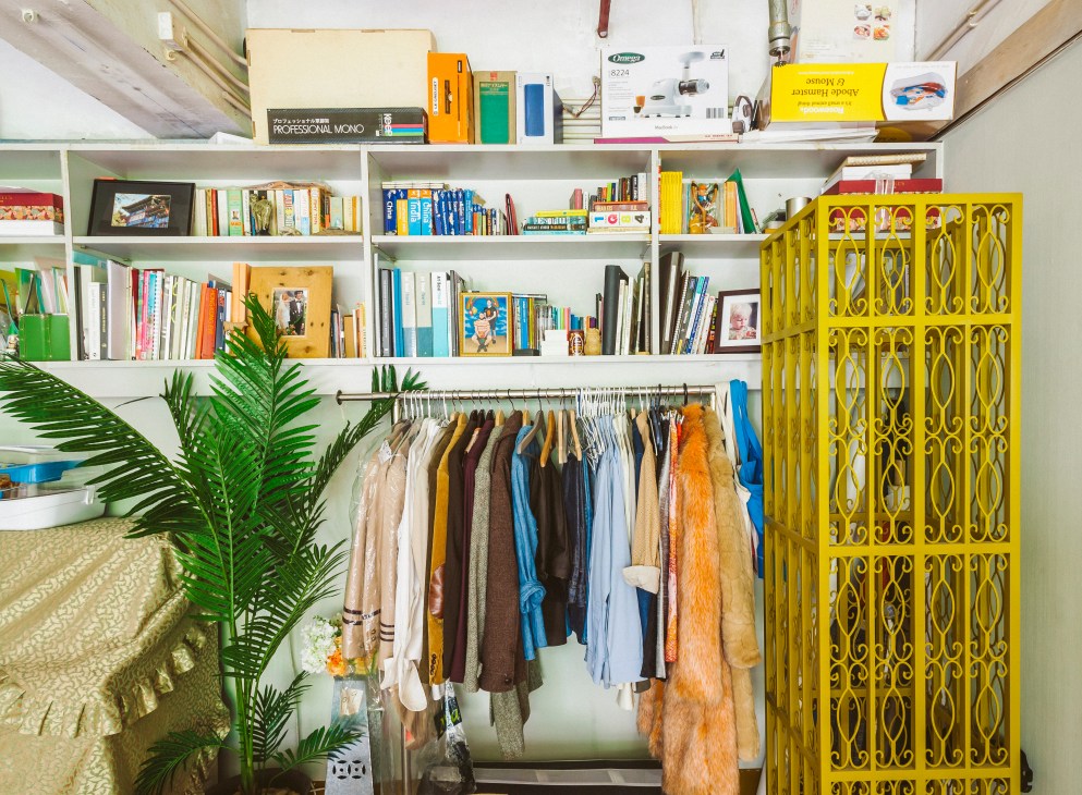 Adrian Wong's studio workspace with white shelving displaying art books, supplies, and clothing on hangers below.
