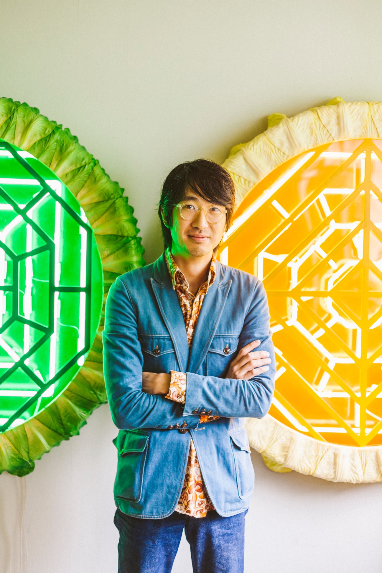 Adrian Wong stands with arms crossed in front of his neon artwork 'Untitled Grate' featuring green and yellow geometric designs.
