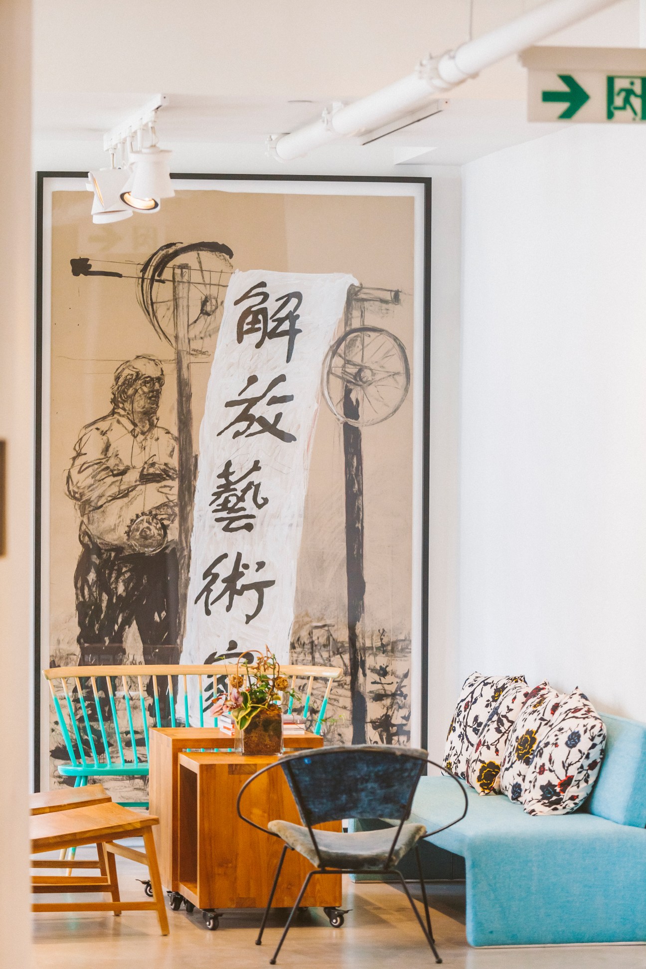 Modern art studio lounge with turquoise chair, potted plant, and large framed artwork featuring Chinese characters and bicycle illustrations on white wall.