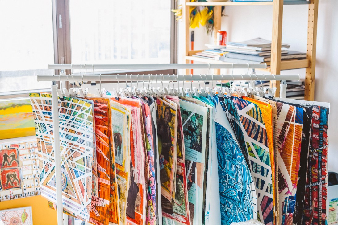 Colorful artwork samples hanging on a white rail in a bright studio space with large windows.