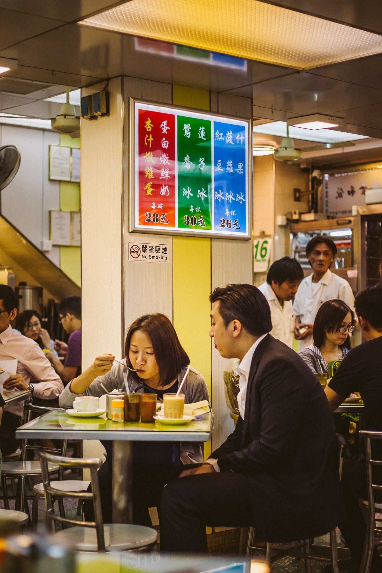 Diners at Australian Dairy Company in Hong Kong, a cha chaan teng restaurant with colorful menu boards displaying dishes and prices in Chinese.