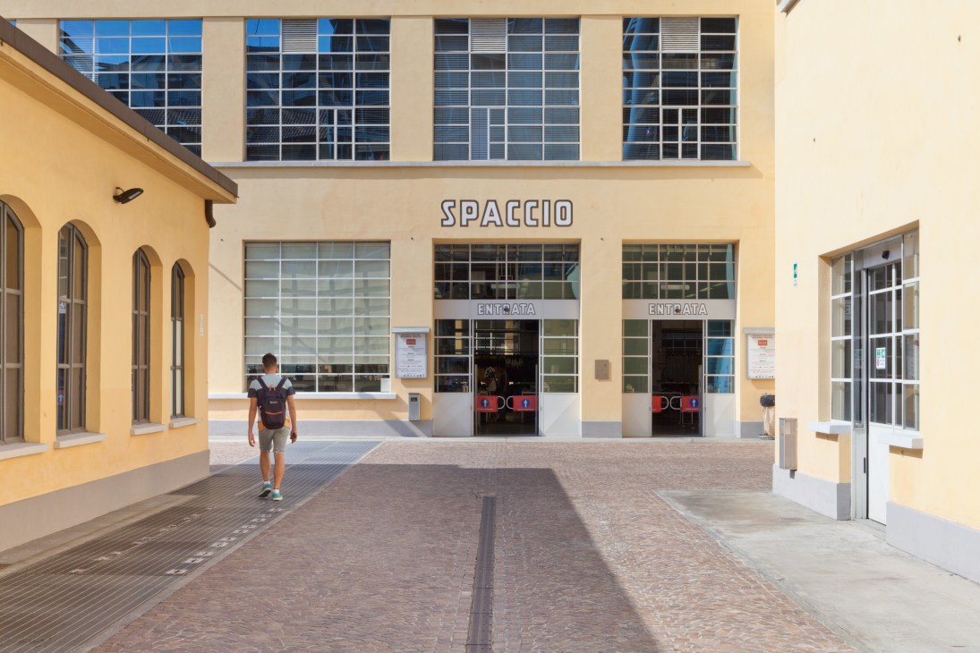 A person walking through a sun-lit courtyard in BasicVillage, a converted factory with yellow ochre walls and large windows marked with 'SPACIO' signage.