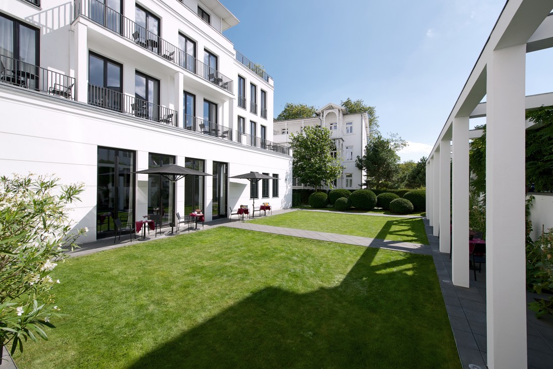 Modern white hotel building with manicured lawn, garden seating area, and Scandinavian-style architecture.