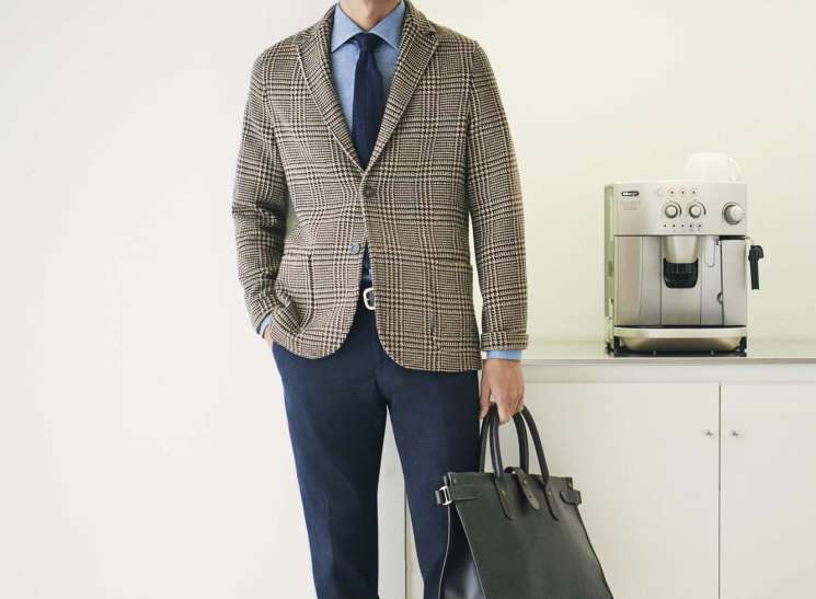 Man in beige checked blazer, blue shirt and navy trousers holding a dark leather bag in a minimalist kitchen setting.