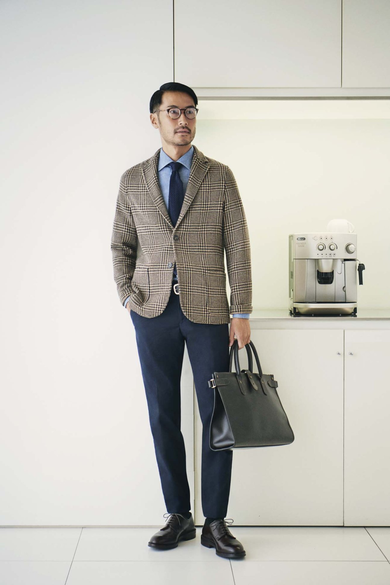 Man in beige checked blazer, blue shirt and navy trousers holding a dark leather bag in a minimalist kitchen setting.