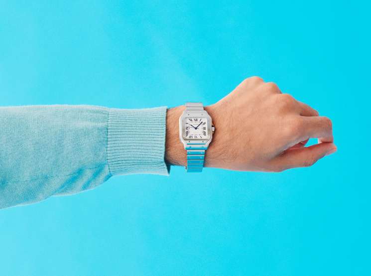 Man's wrist wearing a Cartier watch with a turquoise jumper sleeve against a bright blue background.