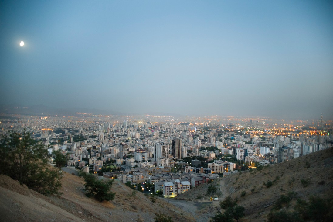 Panoramic view of northern Tehran's sprawling cityscape at dusk with moon visible in blue sky.