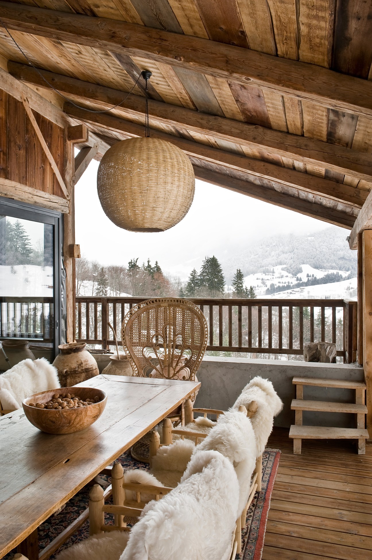 Alpine chalet porch with wooden dining table, woven pendant light, and snowy mountain views.