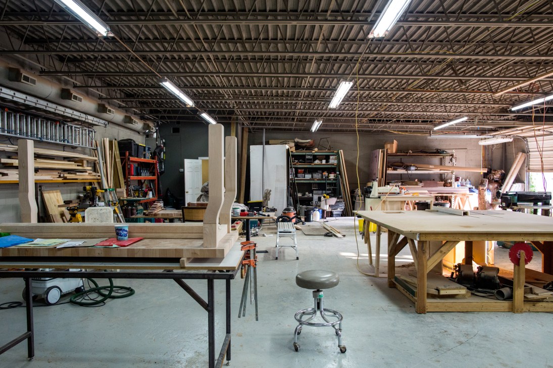 Interior of Fecht Designs woodworking workshop with workbenches, tools, and materials in an industrial space.