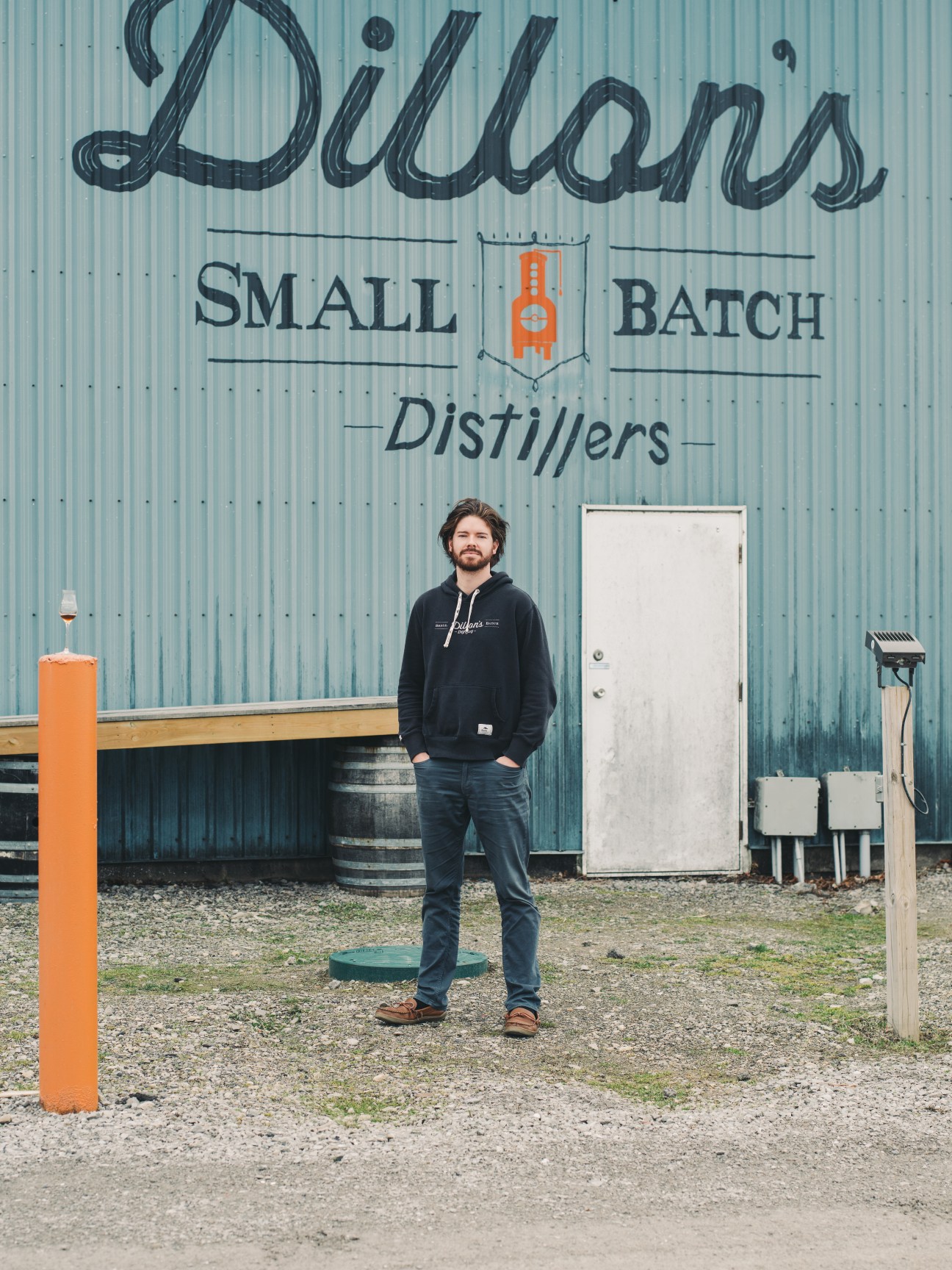 Geoff Dillon stands in front of Dillon's Small Batch Distillers building with corrugated metal walls.