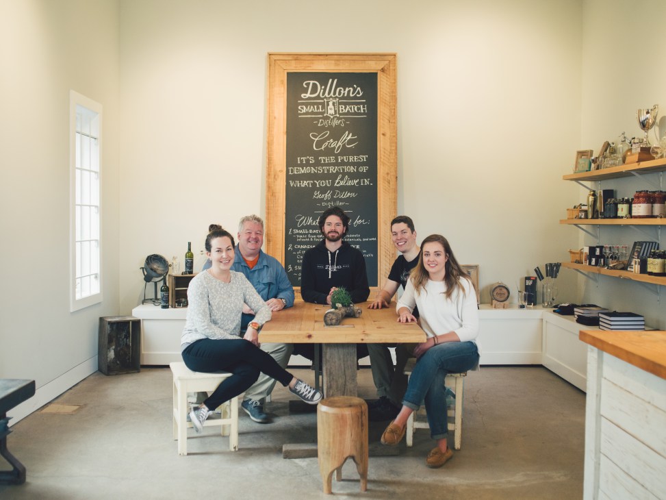 Five members of the Dillon's distillery team pose together at a wooden table in their bright, minimalist tasting room with a chalkboard menu behind them.