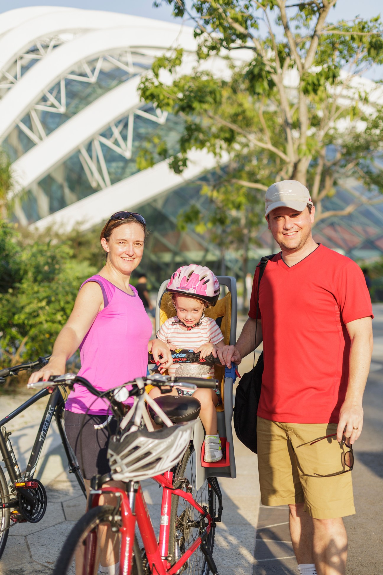 Family with child in bike seat posing with bicycles in front of Gardens by the Bay's white Supertree structures in Singapore.