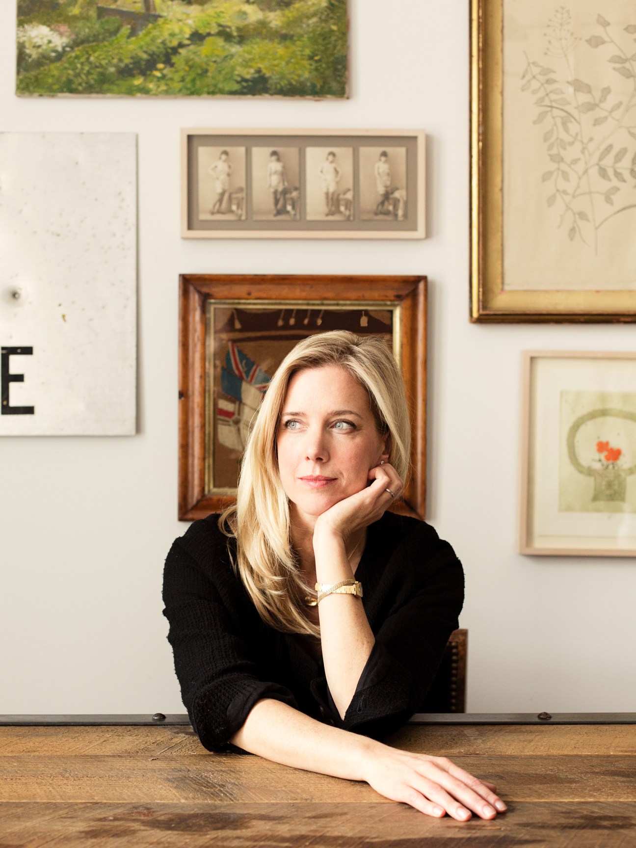 Portrait of Elizabeth Roberts, a designer with blonde hair wearing a black top, seated at a wooden desk with framed artwork on the wall behind her.