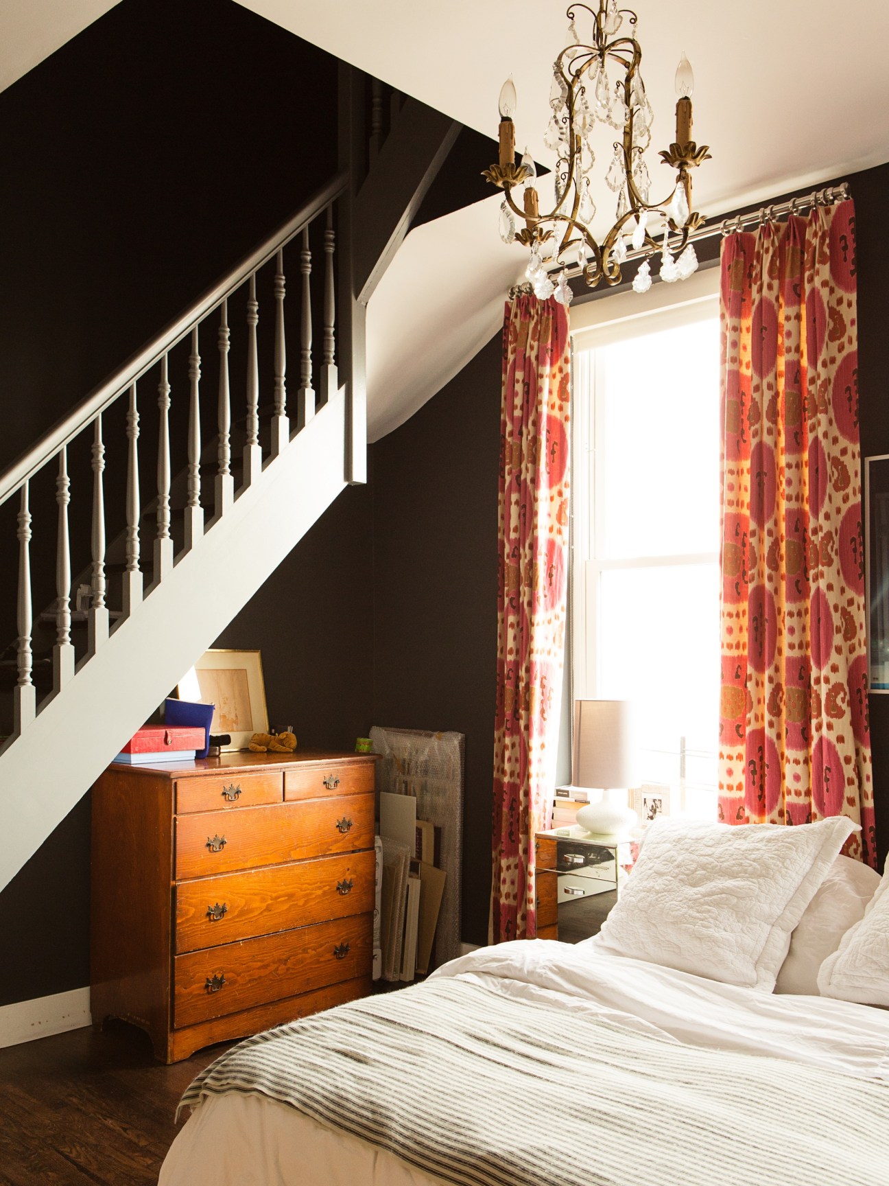 Bedroom with wooden chest of drawers, ornate chandelier, red patterned curtains, and black accent wall beneath white staircase.