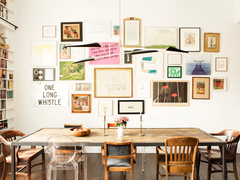 Dining room with wooden table, mismatched vintage chairs, and gallery wall of framed art and text on white walls.