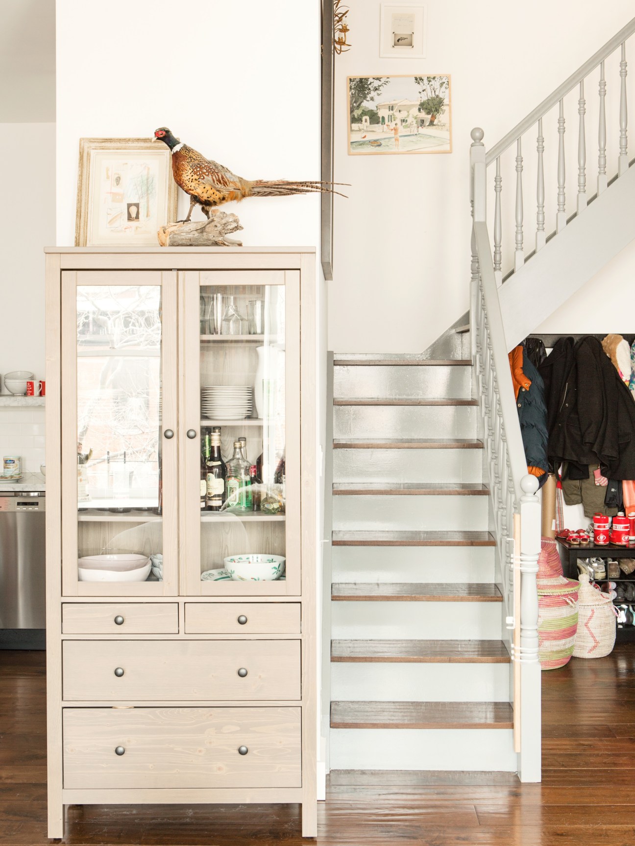 Kitchen cabinet with glass doors displaying dishware and taxidermied pheasant sculpture on top, next to staircase with metal railings.