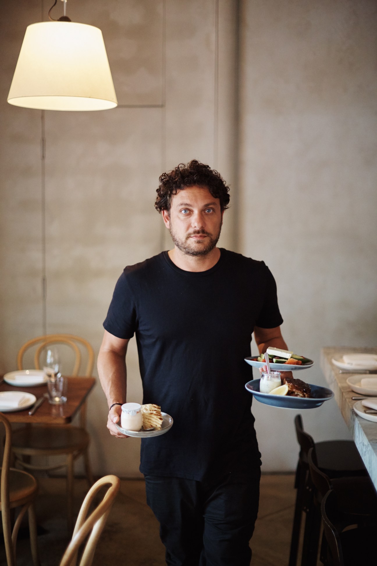 Chef Jonathan Barthlemess holding plates of food in a modern restaurant dining room.