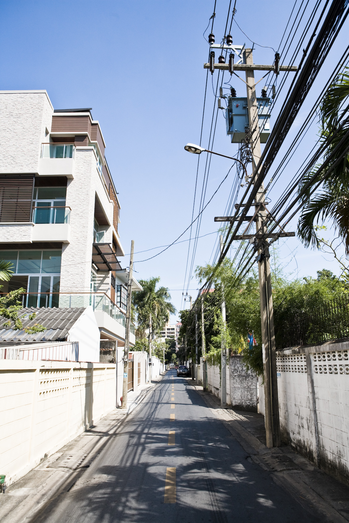 A tree-lined residential street in Ari with a new white apartment building on the left and power lines overhead.