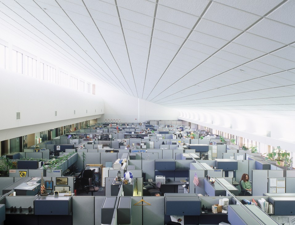 Open-plan office with cubicles and bright natural light from ceiling windows under a gridded metal ceiling.