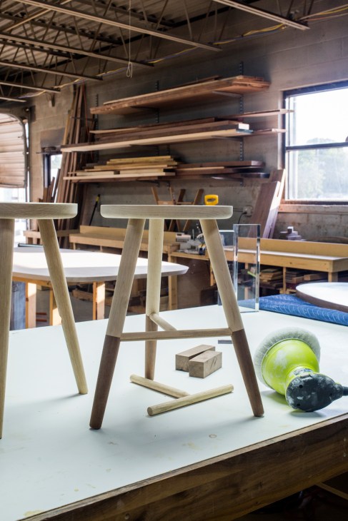 Wooden furniture pieces in progress at Fecht Designs workshop, including a stool with tapered legs and wood shelving with materials in background.