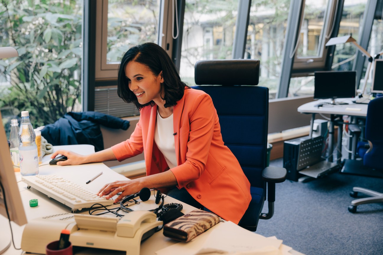 Woman in orange blazer working at desk with keyboard and microphone in modern office with large windows.