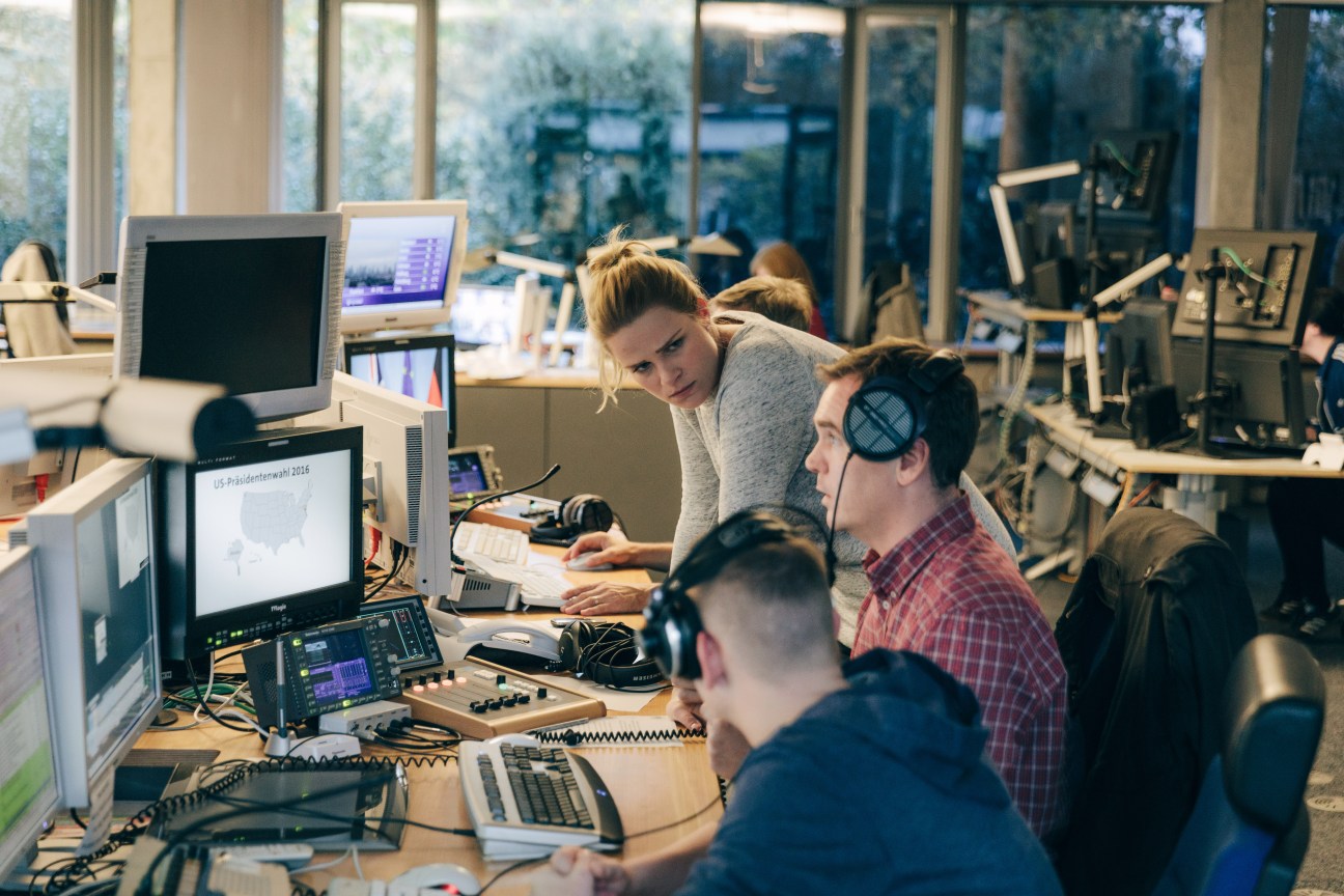 Team members work at desks with computers and equipment in a bright newsroom as deadline approaches.