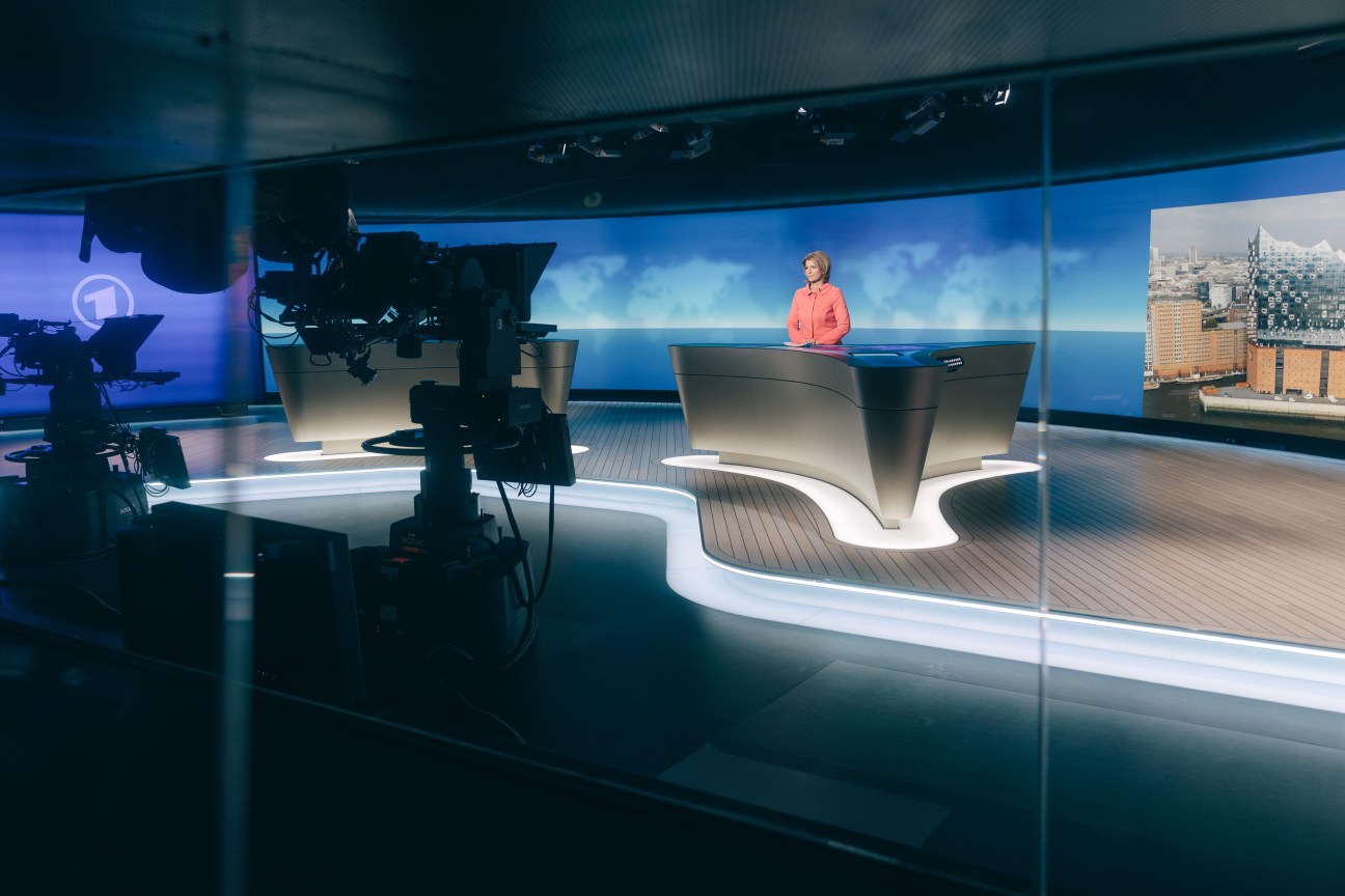 Tagesschau presenter in pink jacket at news desk in modern Hamburg studio with cameras and cityscape backdrop.