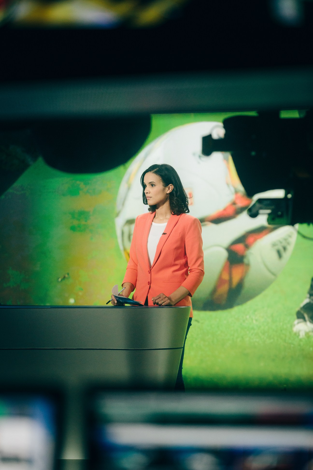 Tagesschau24 presenter Charlotte Maihoff stands at the news desk in a coral blazer, with a screen displaying news graphics behind her.