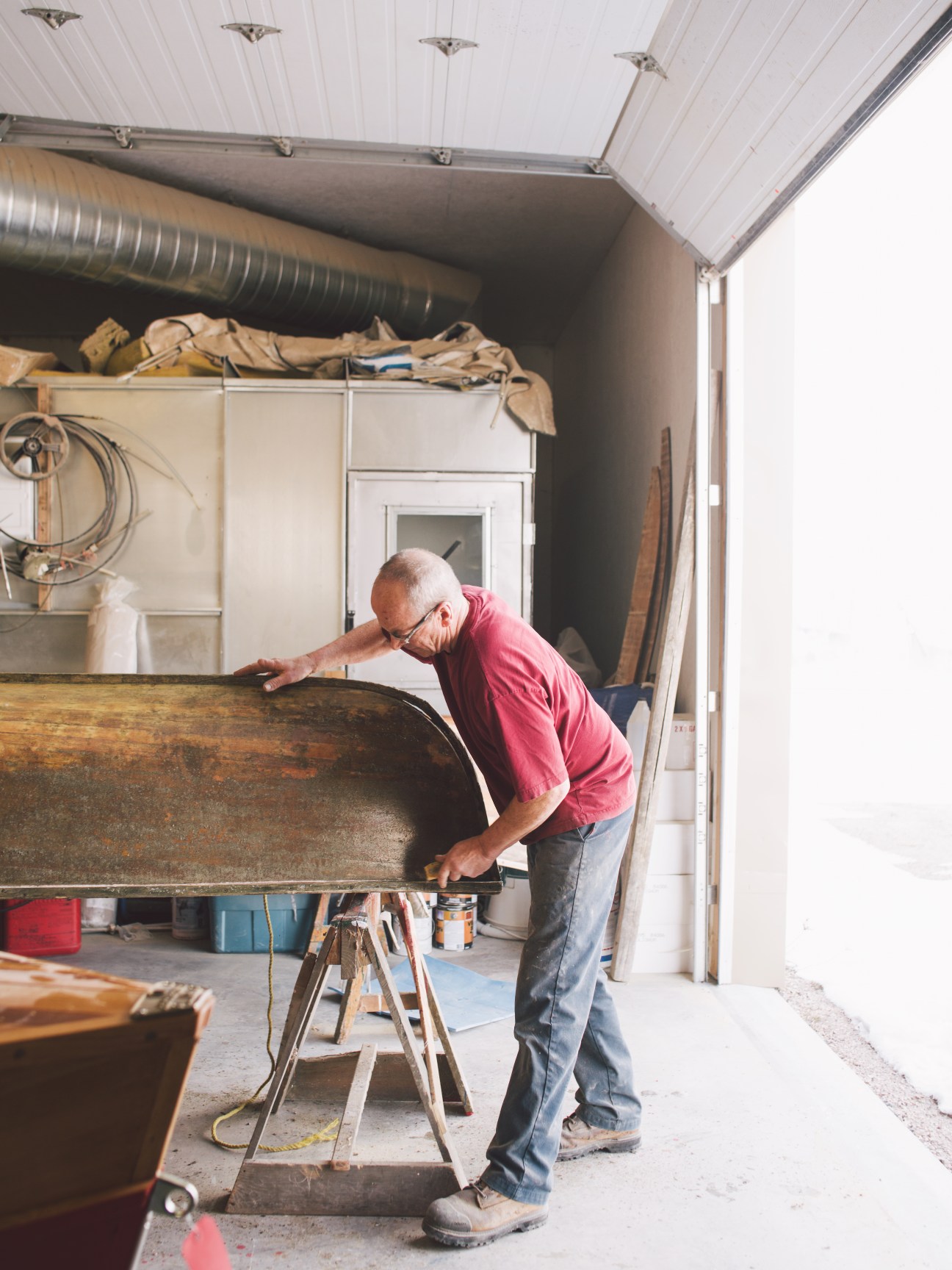Man in pink shirt scraping dirt off an old wooden canoe inside a workshop garage.