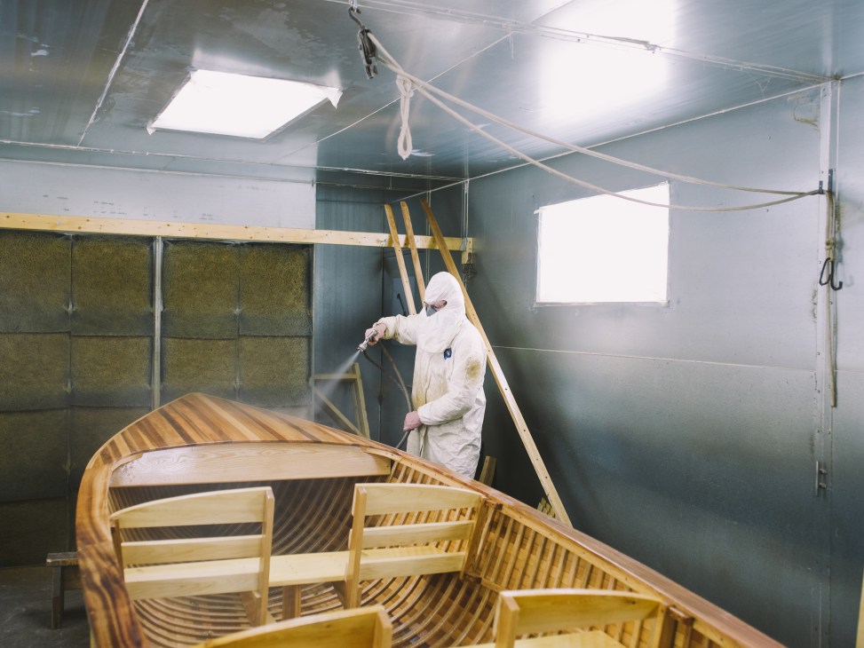 Worker in white protective gear spraying varnish on a wooden rowboat in a workshop.