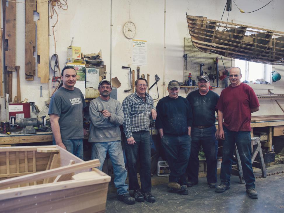 Six men standing in a wooden boat workshop with tools and boat hulls visible.