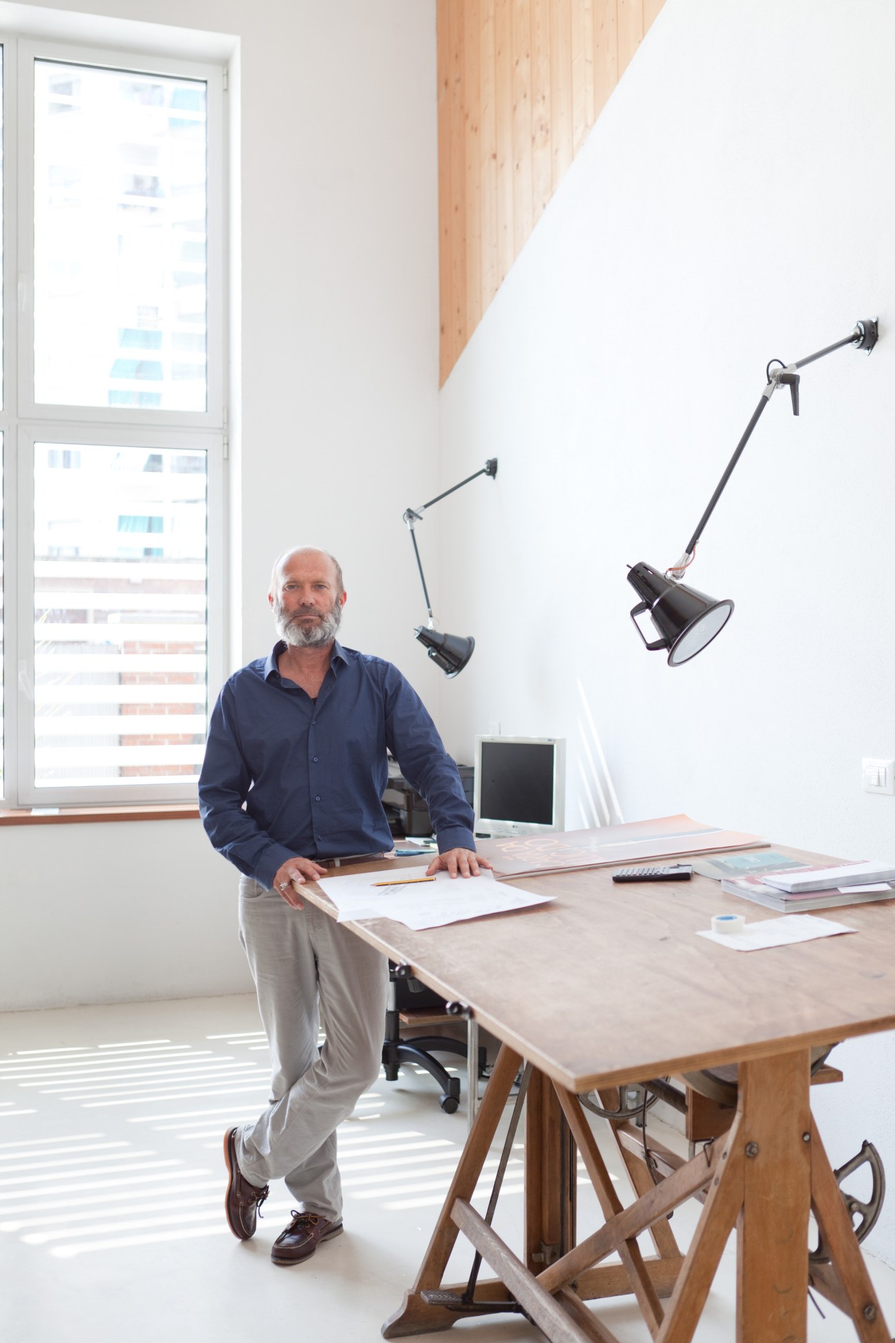 Architect Maurizio Zucca standing at a wooden desk in a bright studio space with industrial lamps and large windows overlooking Turin.