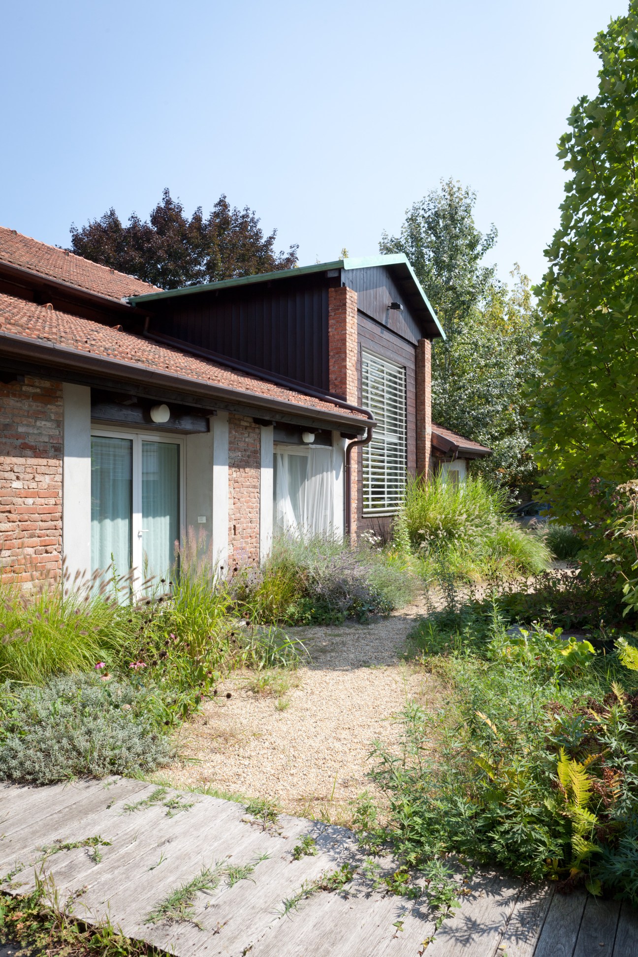Modern brick residential building with black metal accents and large windows surrounded by mature greenery and gravel pathways.