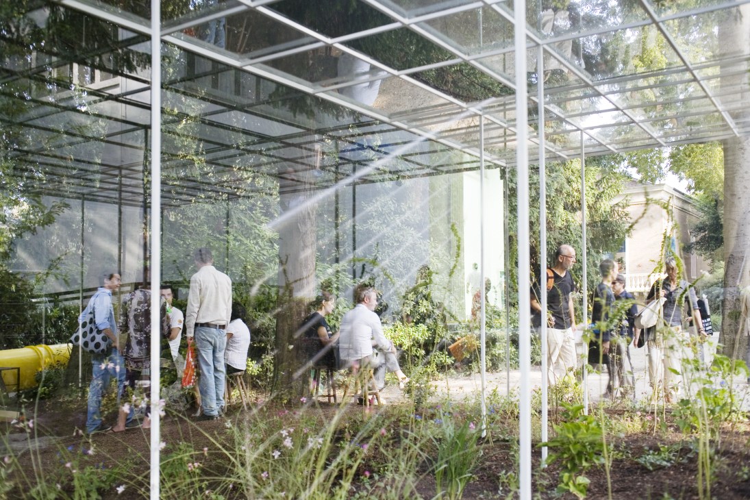 Visitors inside Japan's Venice Architecture Biennale pavilion with glass walls, metal framework, and lush vegetation.