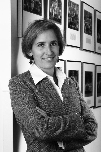 Black and white portrait of Xenia Dormandy, a woman with short dark hair wearing a cardigan and white collar shirt, standing with crossed arms in front of framed photographs on a wall.