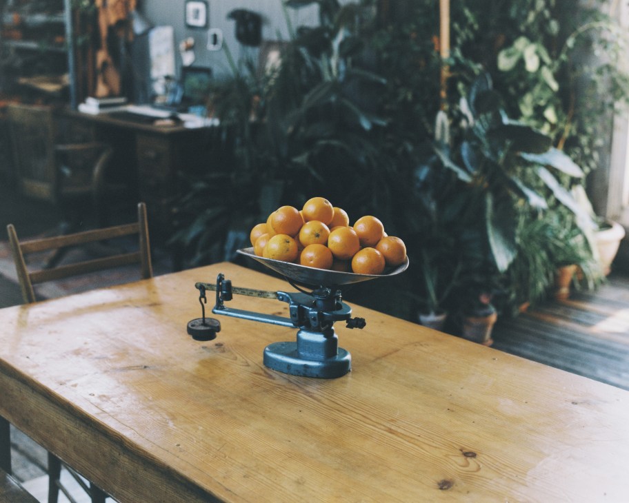Fresh oranges in a bowl on a wooden table with vintage scales and potted plants visible in the background.