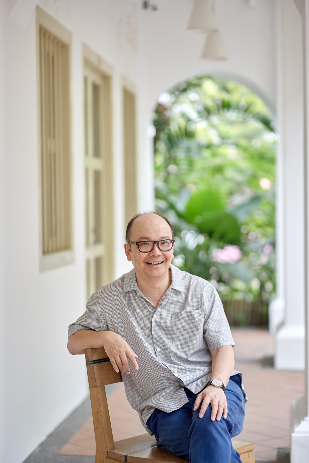 Kay Ngee Tan crouching on a wooden chair in a white corridor with an arched garden view