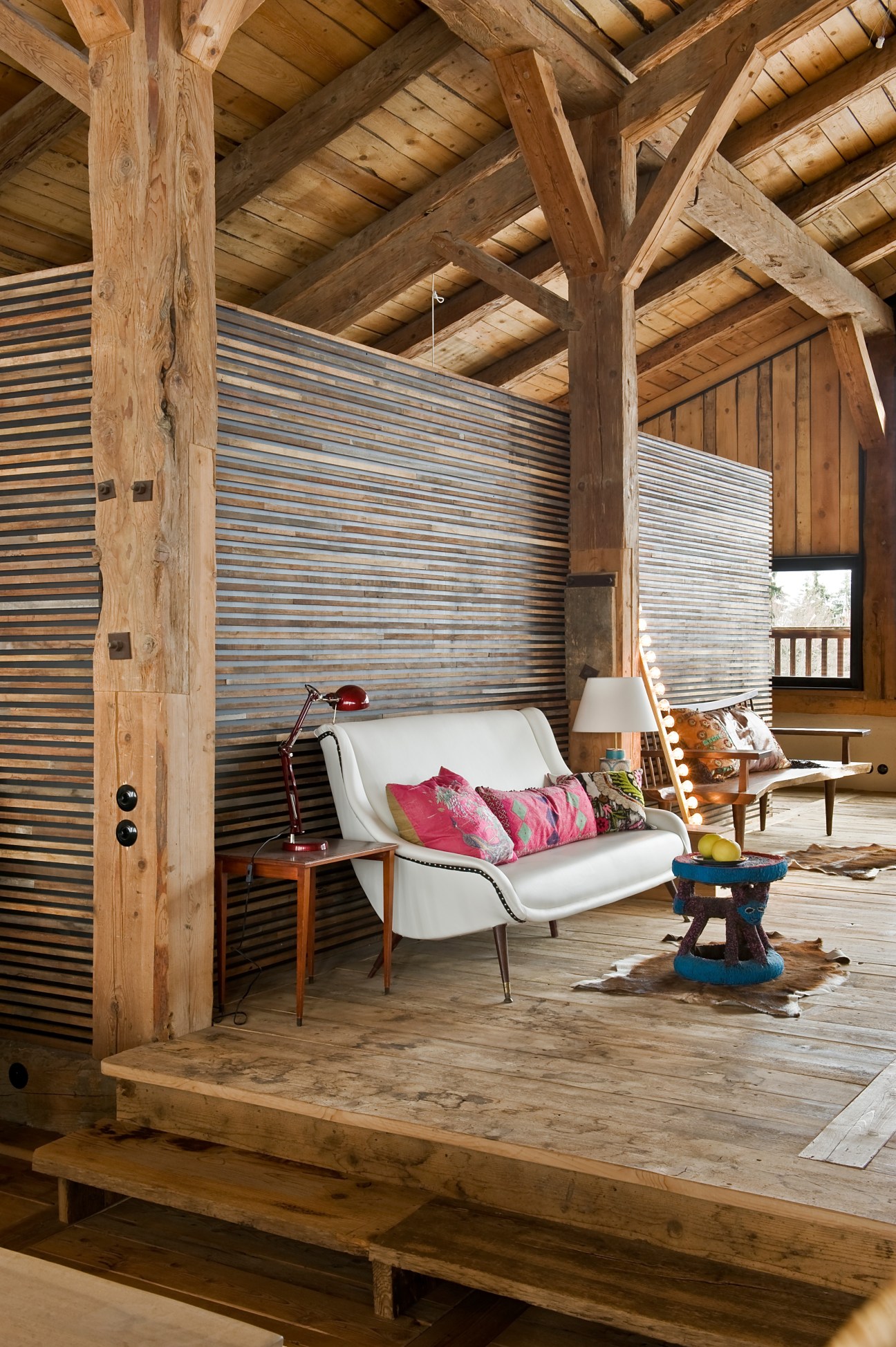 Alpine chalet interior with white mid-century lounge chair, vintage cushions, exposed wooden beams, and corrugated metal wall panels.
