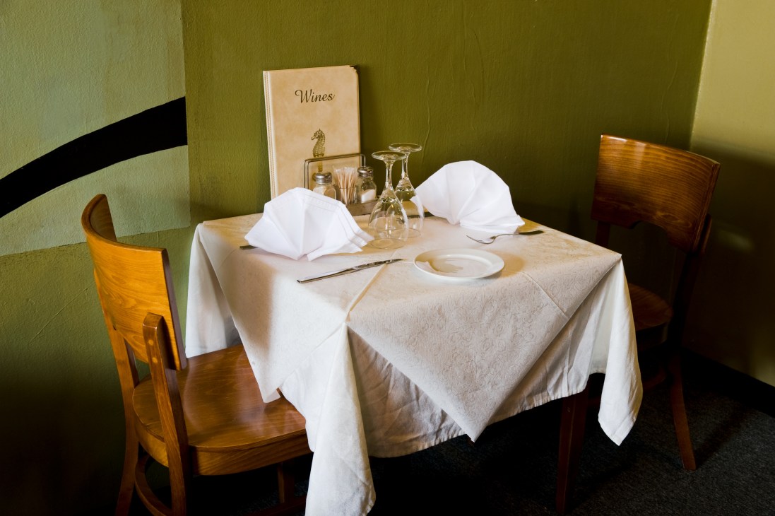 Intimate dining table set for two with white napkins, wine glasses, and a wines menu in a green-walled restaurant.