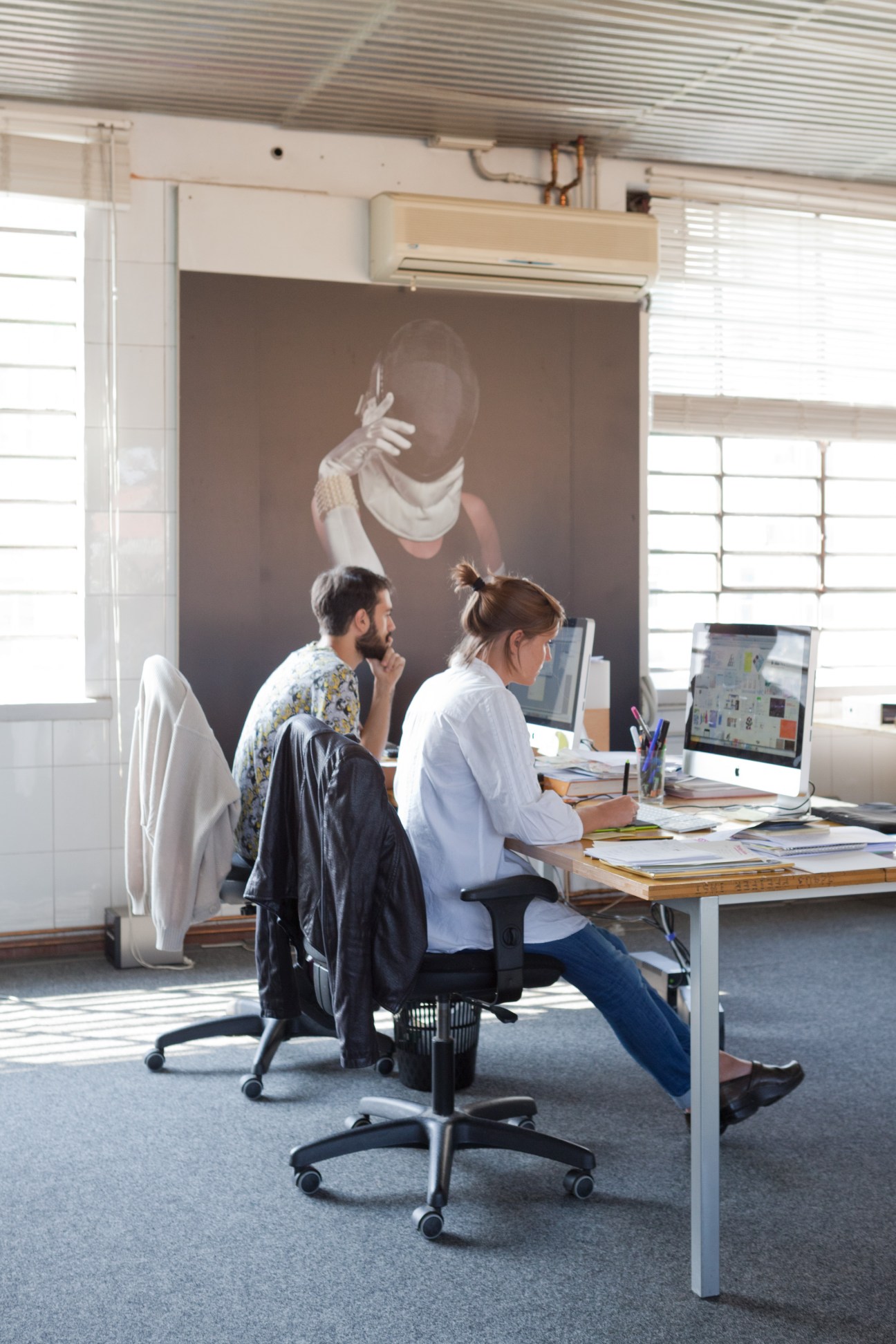 Two people work at a desk in a bright industrial loft space with large windows and artwork on the wall.
