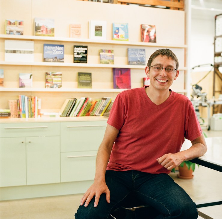 Man in red shirt smiling at camera in front of wooden shelving with books and plant.