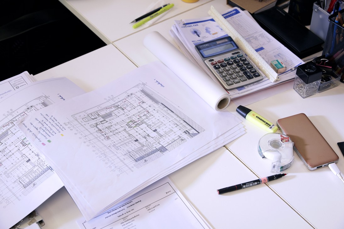 Architectural floor plans and blueprints spread across a white desk with design tools including a calculator, highlighter, and smartphone.