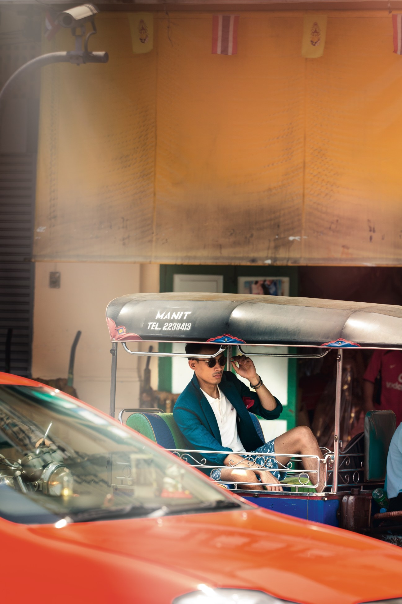 Man in blue jacket and sunglasses riding a Bangkok tuk-tuk with orange awning.